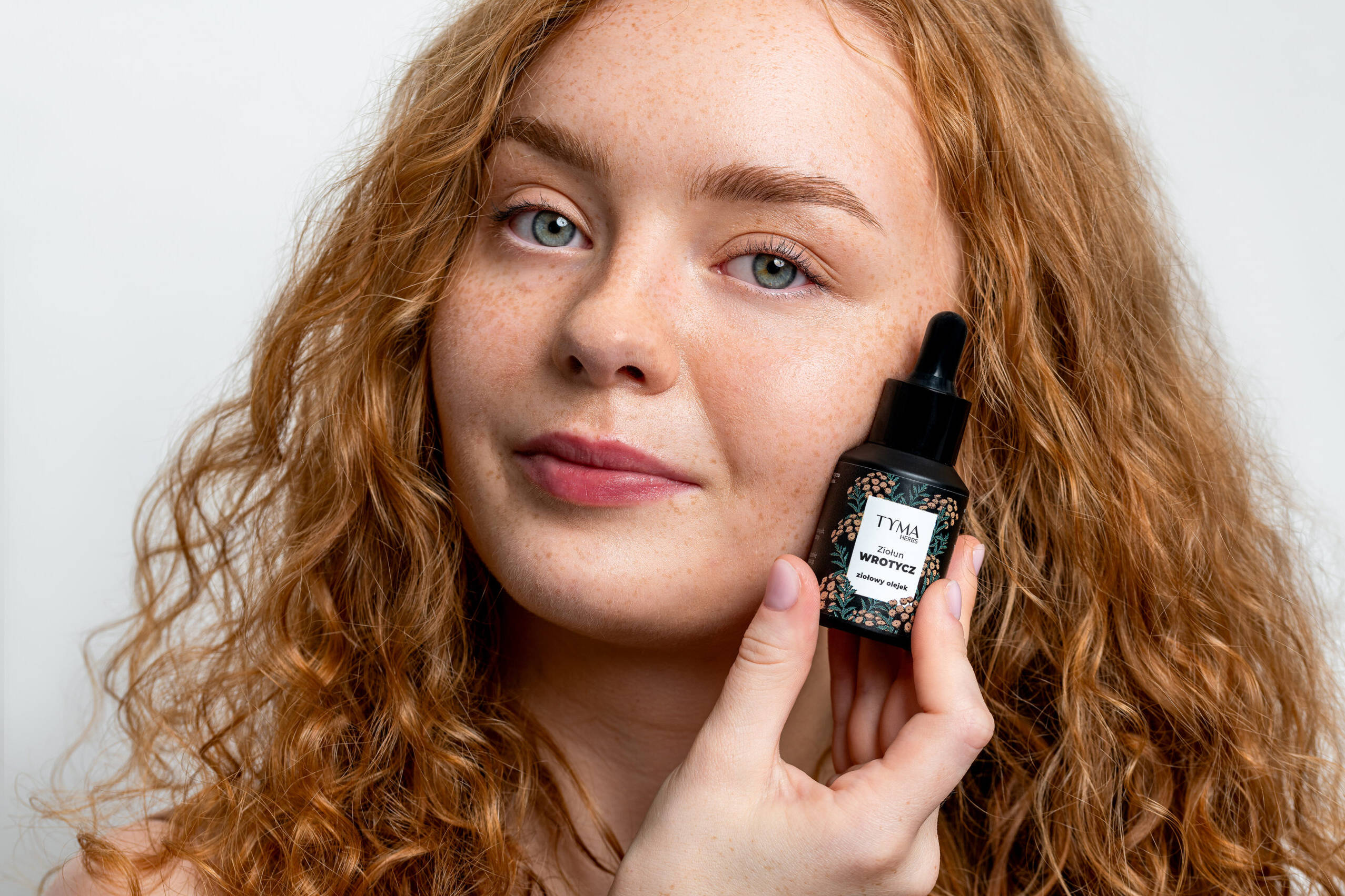 Modelka z produktem marki Tyma Herbs, z branży ziołowych kosmetyk&oacute;w. Rudowłosa kobieta, sesja studyjna.